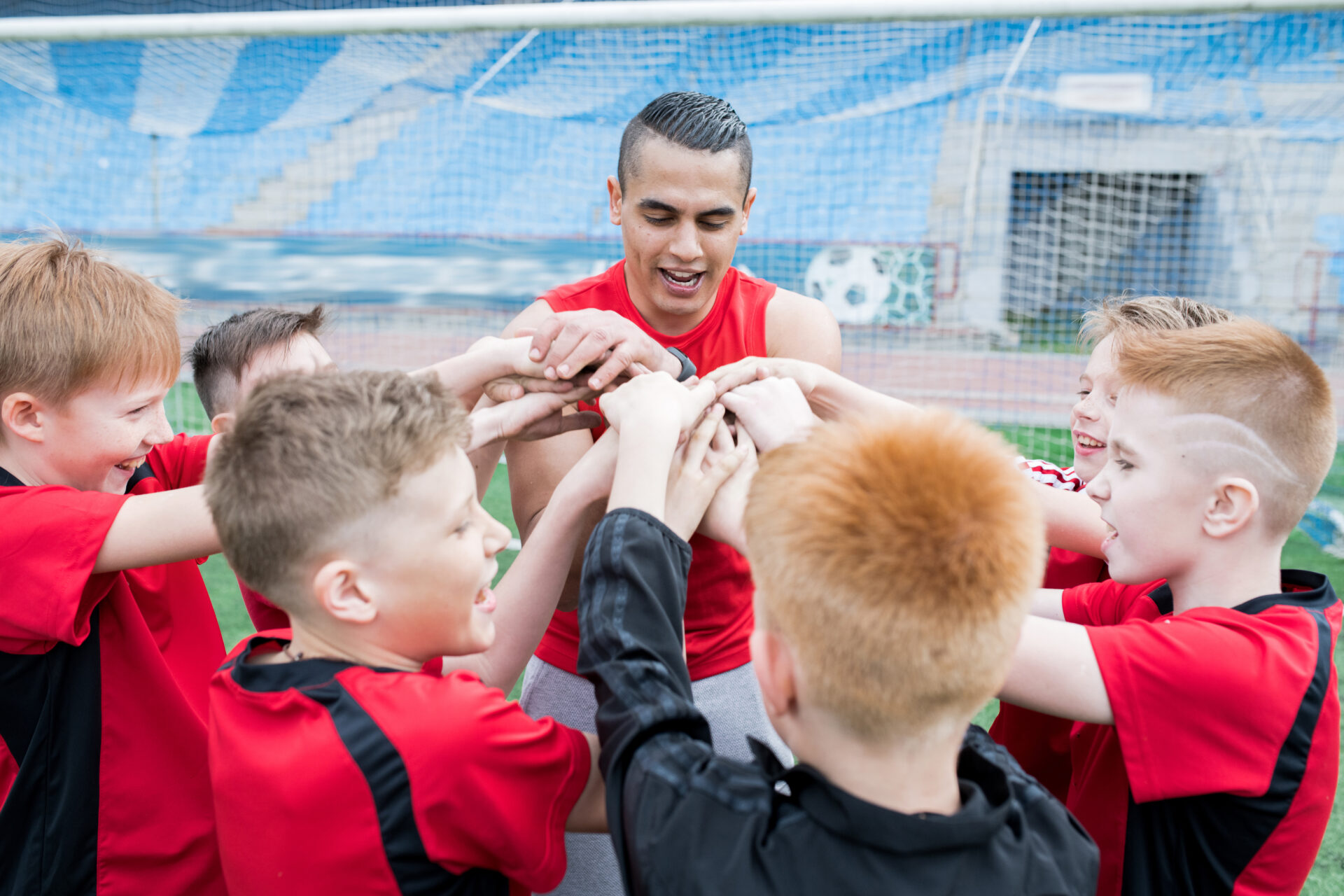 Portrait of junior football team stacking hands during motivational pep talk before match in outdoor stadium, focus on young coach